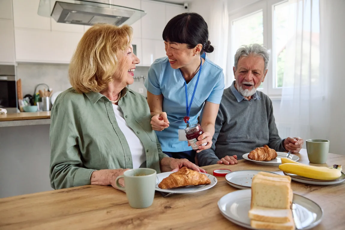 Caregiver assisting older adults with breakfast in a home-style assisted living kitchen.